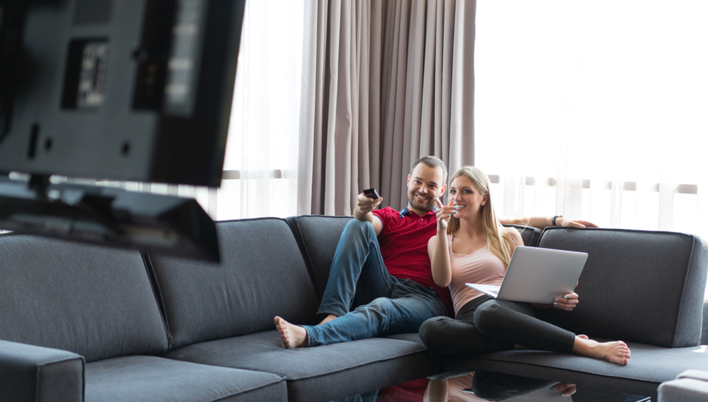 A man and woman sitting an a couch, watching a tv and using a laptop.